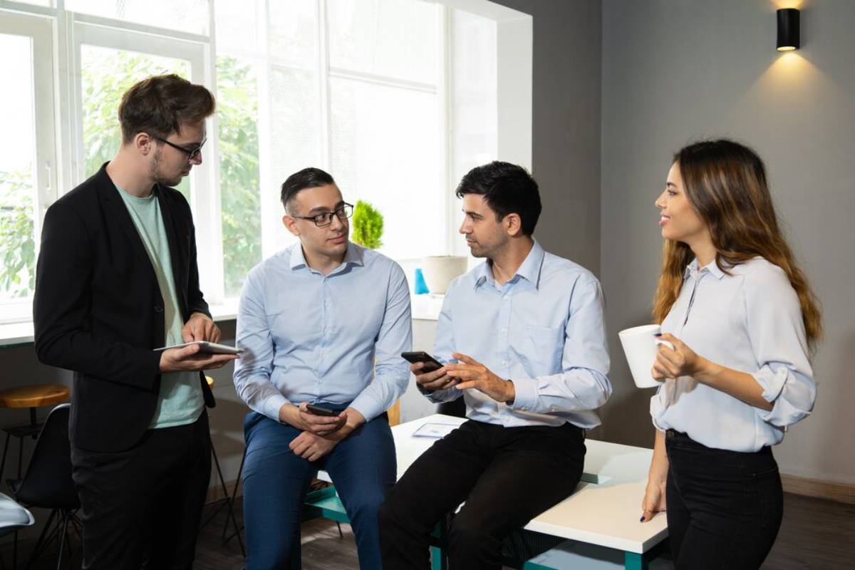 Four professionals engaging in a conversation inside a modern office, holding mobile devices and coffee cups.