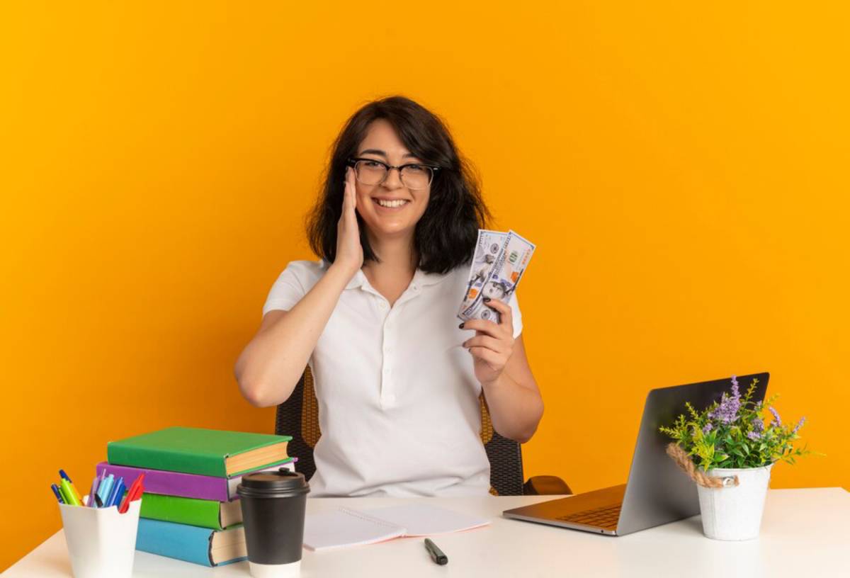 Happy woman holding money, sitting at a desk with books, a laptop, and a coffee cup, against a yellow background.