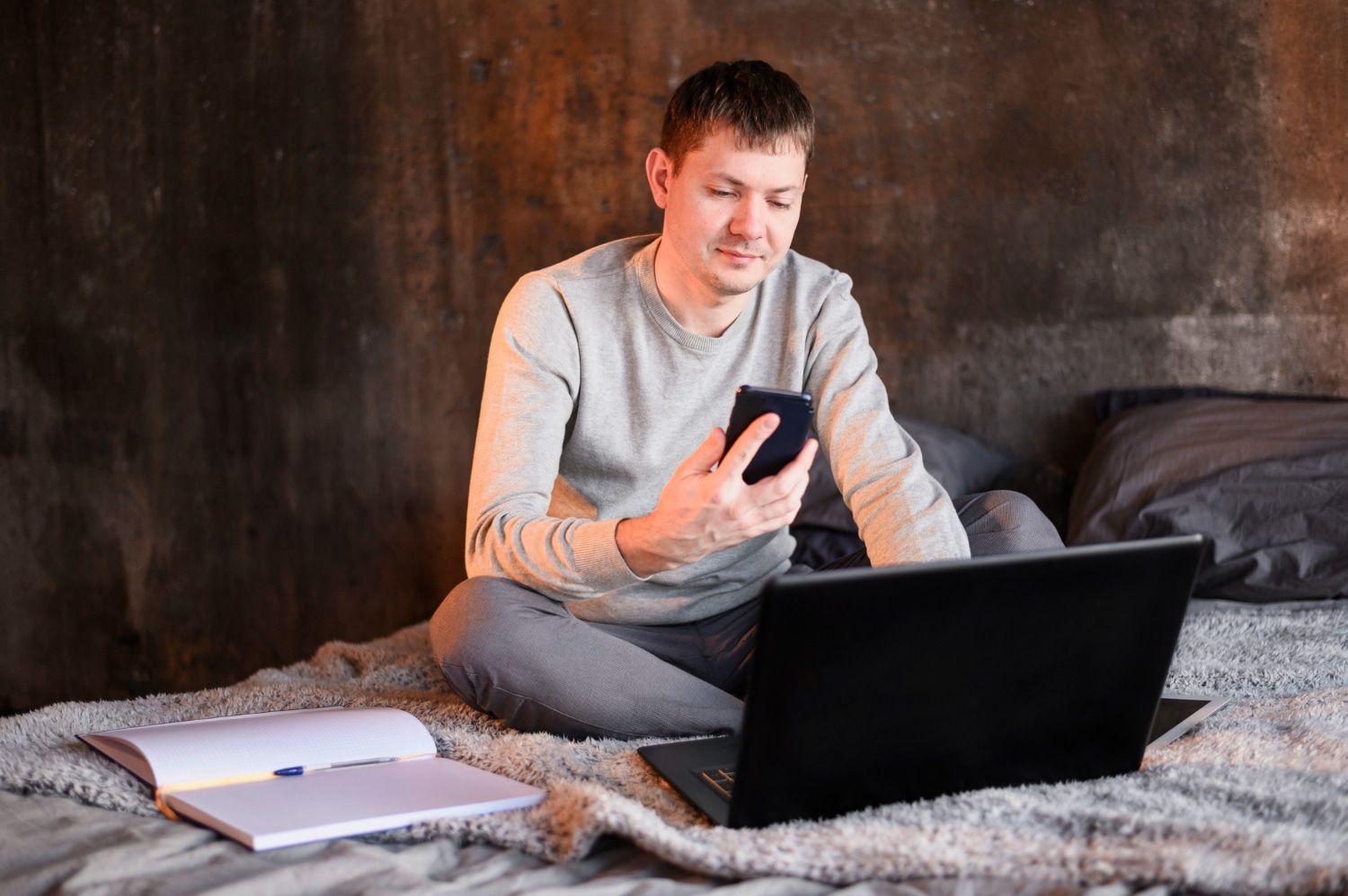 Man sitting on a blanket using smartphone and laptop in a rustic room.