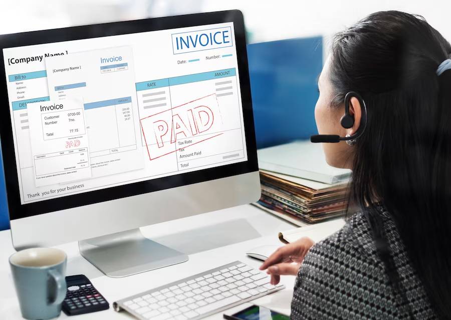 Woman with headset views a digital PAID invoice on computer screen in office setting.