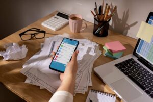 A person using a smartphone with a task management app at a cluttered office desk.