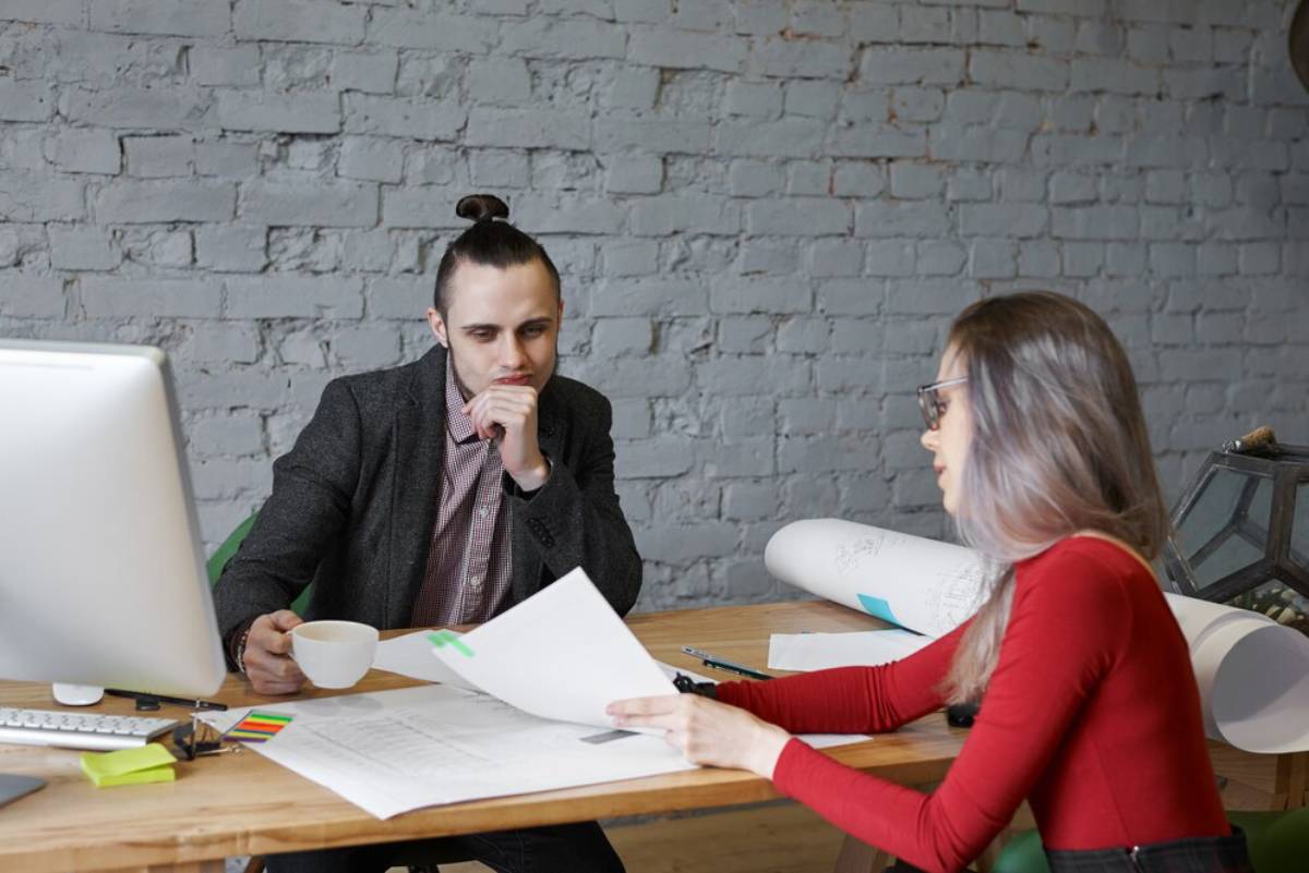 Two professionals, a man and a woman, discussing paperwork at a modern office table with a brick wall background.
