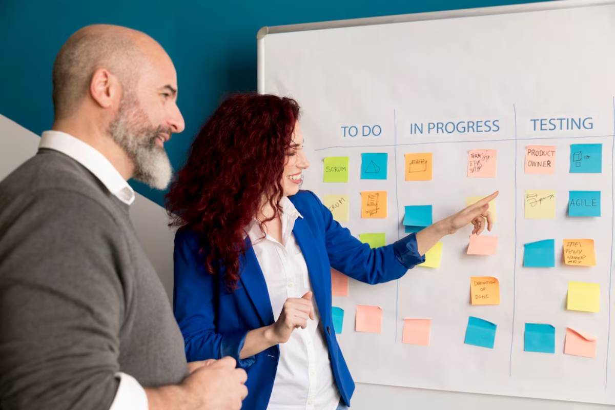 Two professionals discussing workflow on a whiteboard with colored sticky notes in a bright office.