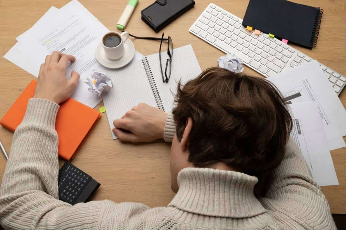 A person resting their head on a desk cluttered with papers, a notebook, a coffee cup, and crumpled paper, indicating stress or exhaustion.