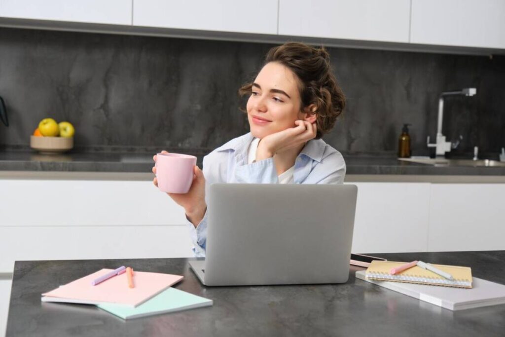 A person sits at a kitchen table with a laptop, holding a pink mug, surrounded by stationery and a bowl of fruit.