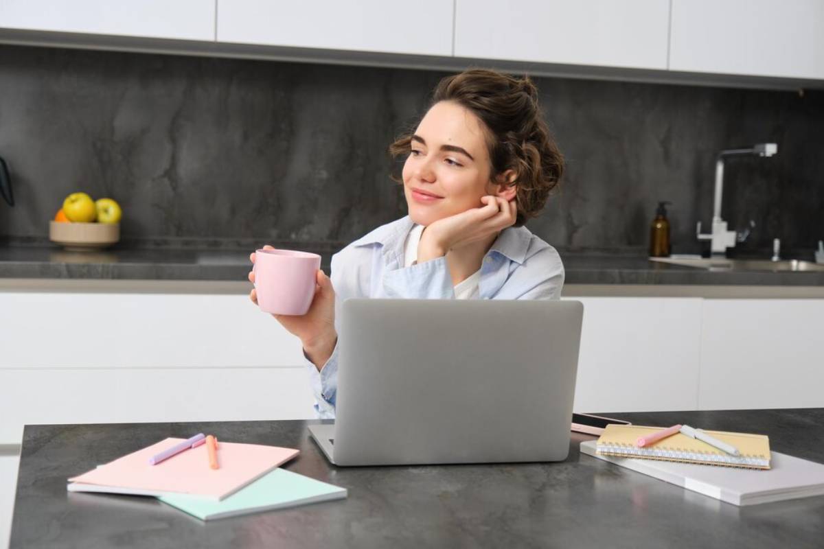 A person sits at a kitchen table with a laptop, holding a pink mug, surrounded by stationery and a bowl of fruit.