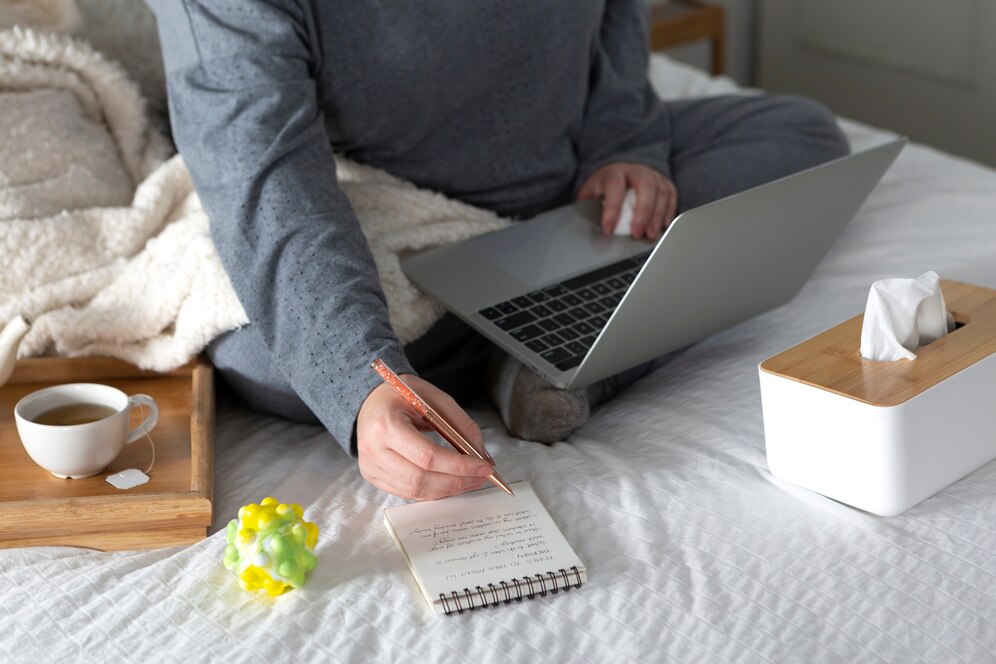 A person in cozy attire sits on a bed, writing notes with a pen while using a laptop, surrounded by tea, a toy, and tissues.