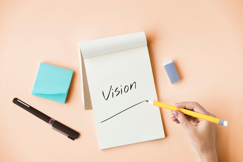 Hand writing the word Vision on a notepad with a pencil, office supplies nearby on a peach background.