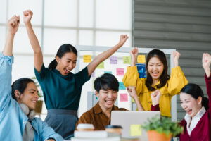 A group of young Asian professionals joyfully celebrates a success at work with laptops and colorful sticky notes around them.