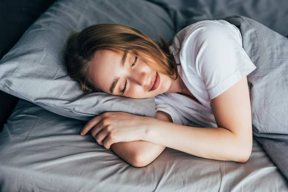 Young woman sleeping peacefully on grey bedding, wearing a white T-shirt.