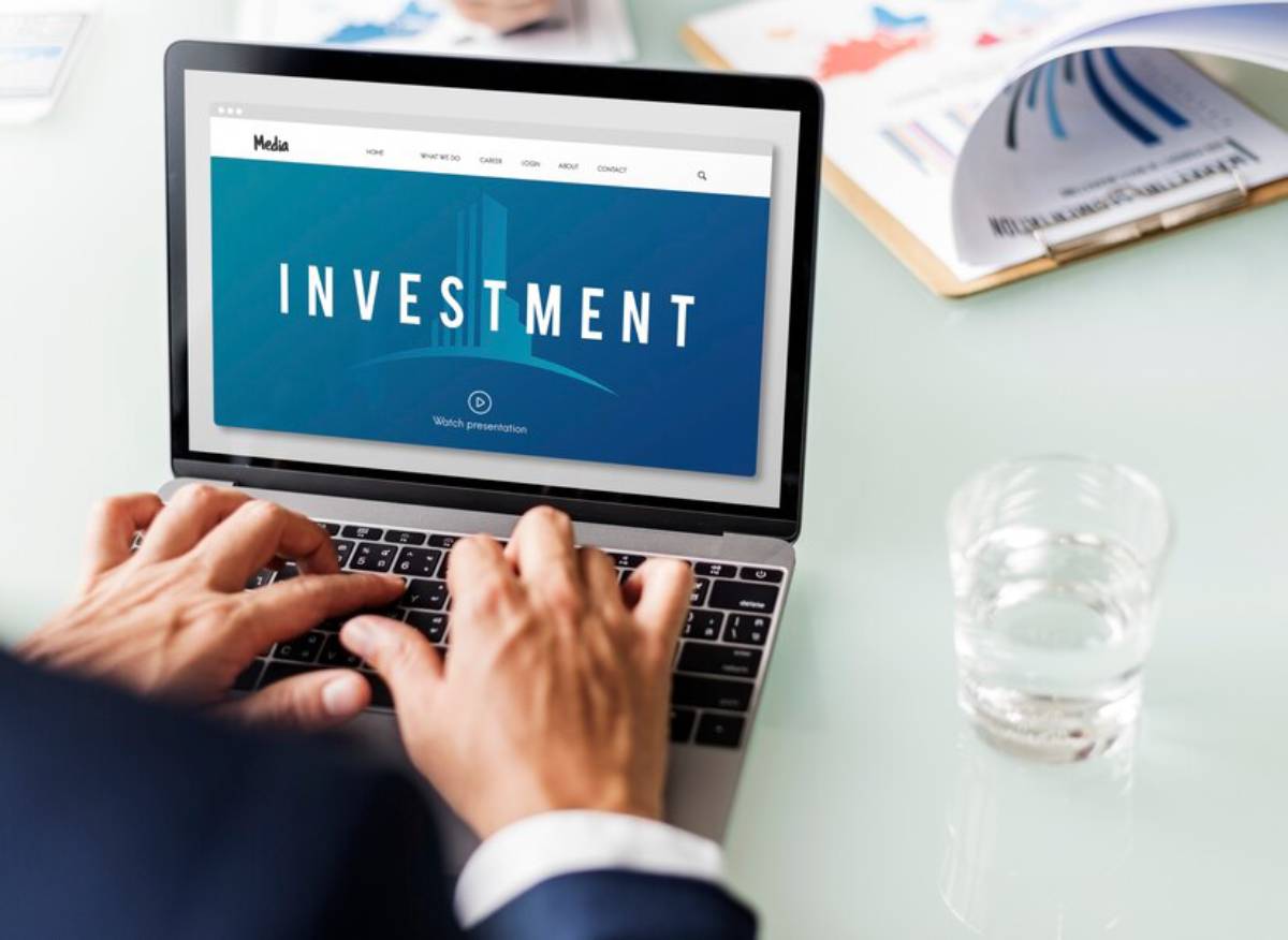 Person using a laptop displaying an investment presentation, with a water glass and financial documents on the table.