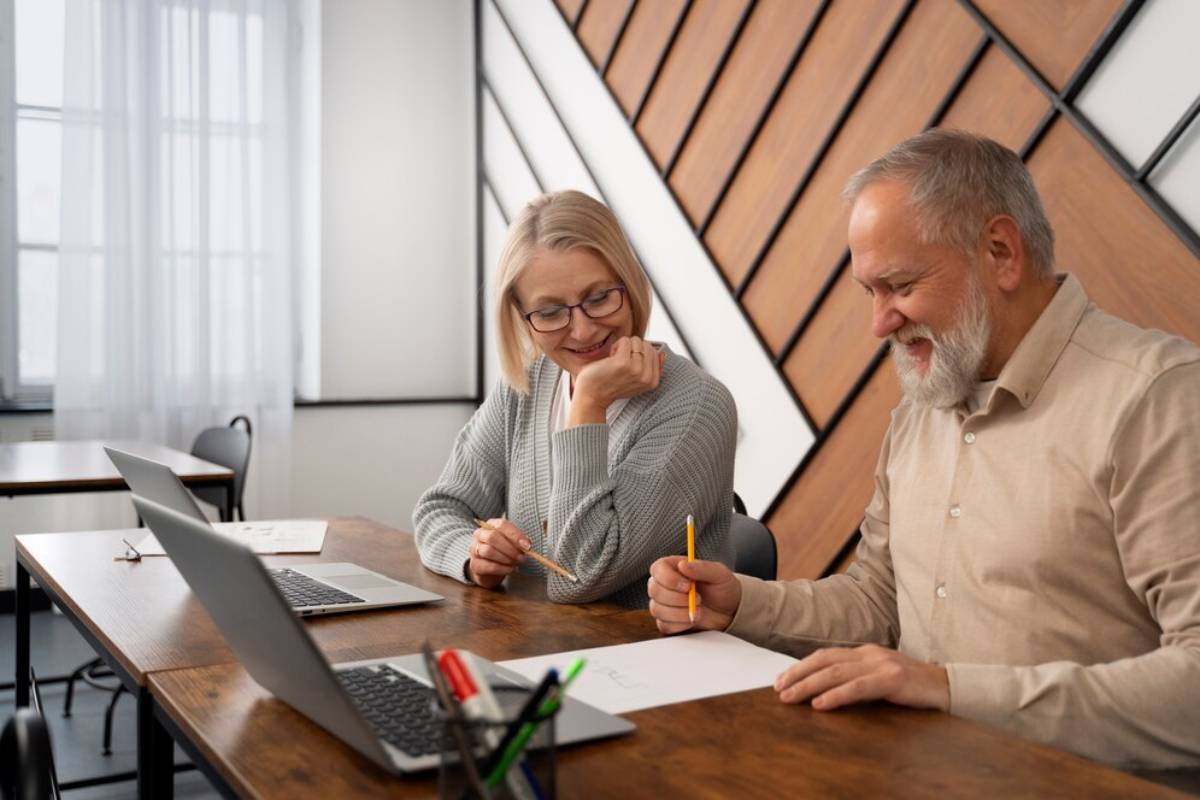 Two senior professionals collaborating over paperwork in a modern office, using laptops and discussing content with smiles.