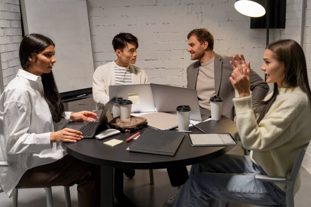 Four colleagues discussing work around a table with laptops and coffee cups in a modern office.