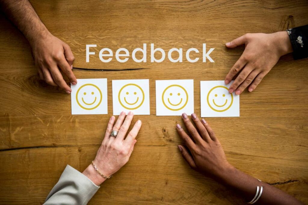 Four diverse hands arranging smiley face cards on a wooden table labeled Feedback.