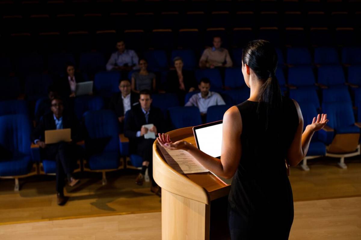 Woman presenting at a lectern in a lecture hall with an audience of attentive professionals.