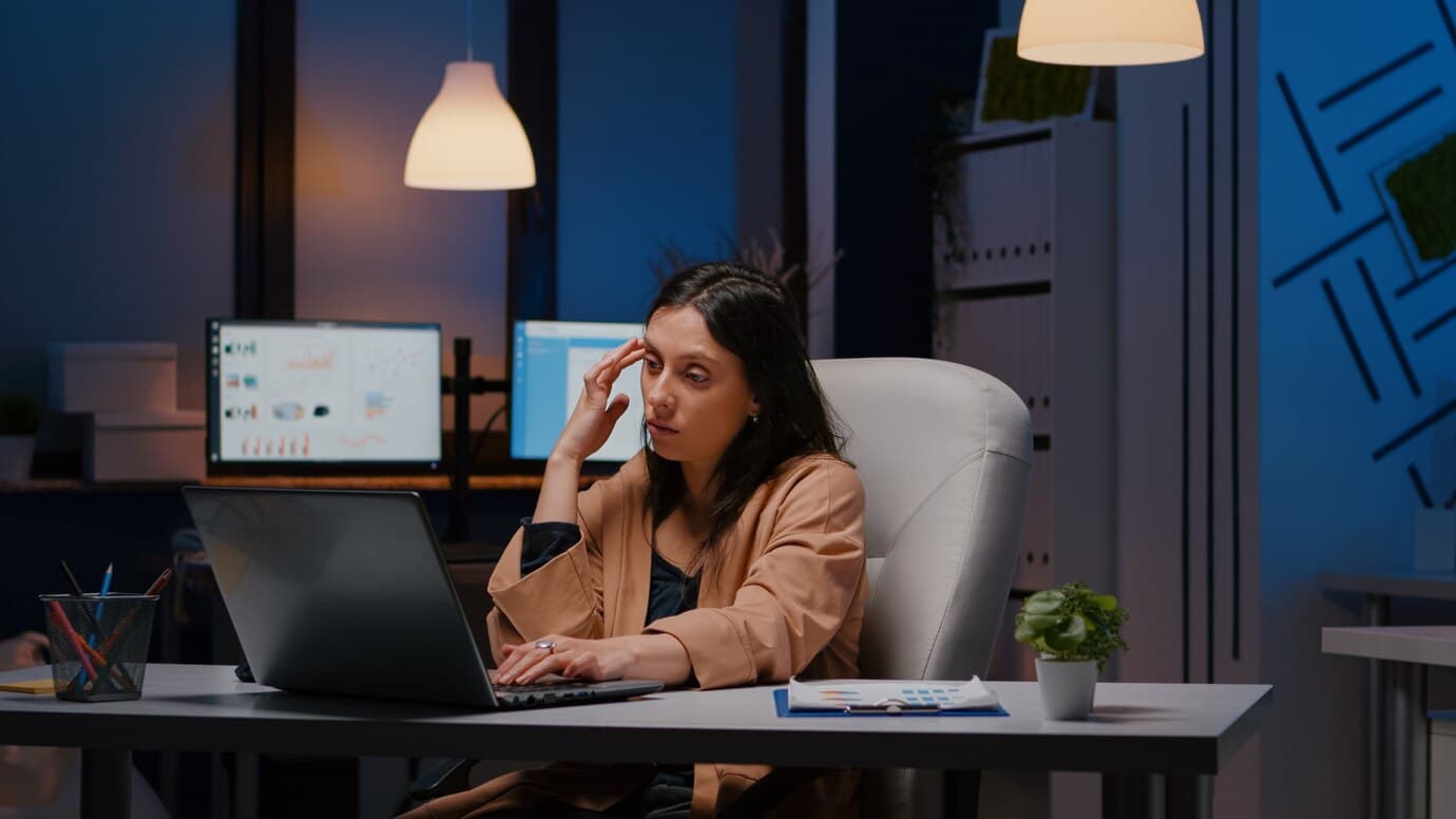 Woman looking tired while working late at office desk with laptops and monitors.