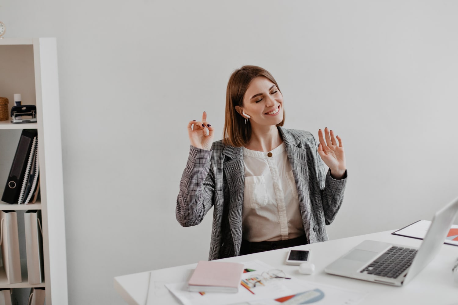 Woman in a plaid blazer joyfully dancing at a tidy office desk with a laptop and papers.