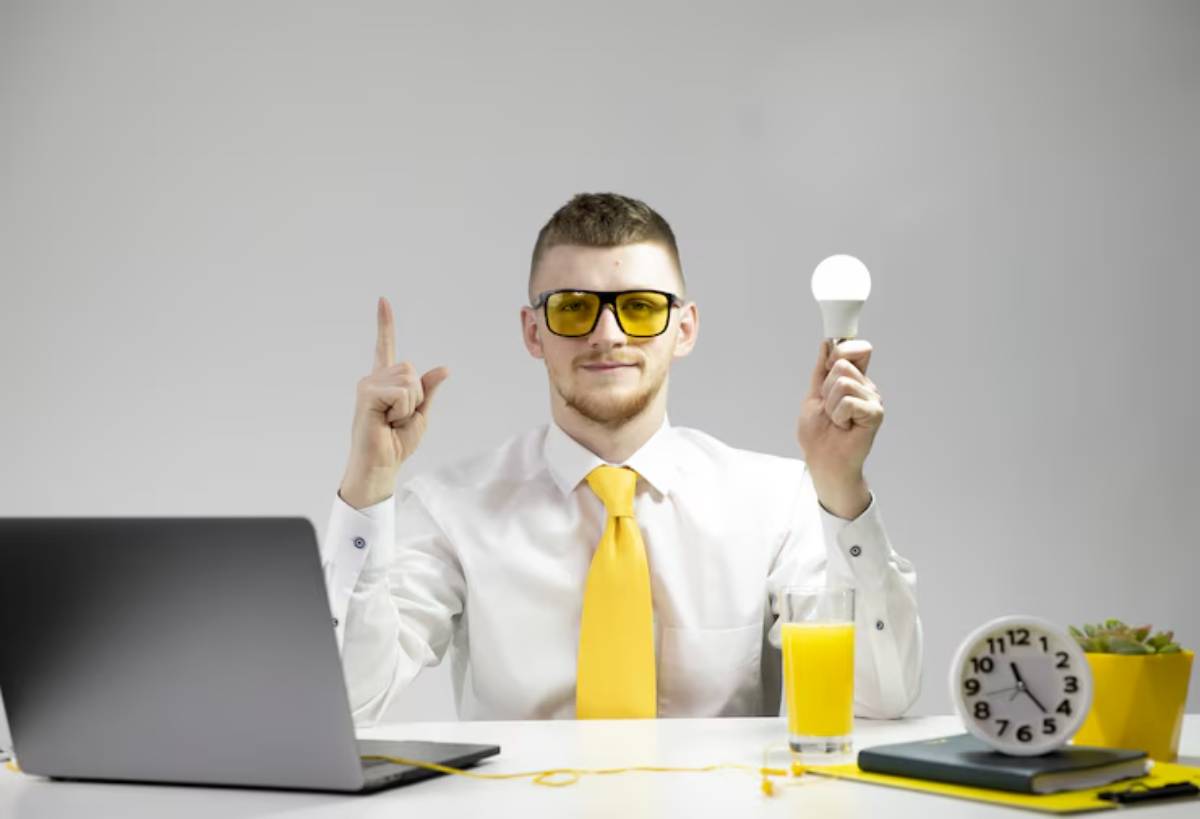 A man in a white shirt and yellow tie holds a light bulb in one hand symbolizing creativity and points upwards with the other, sitting at a desk with a laptop.