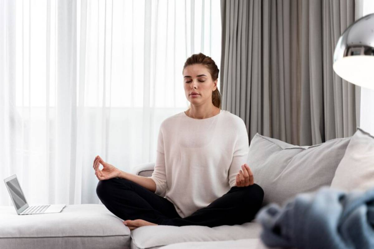 A person in a white sweater meditates on a couch, surrounded by large windows and soft curtains, with a laptop nearby.