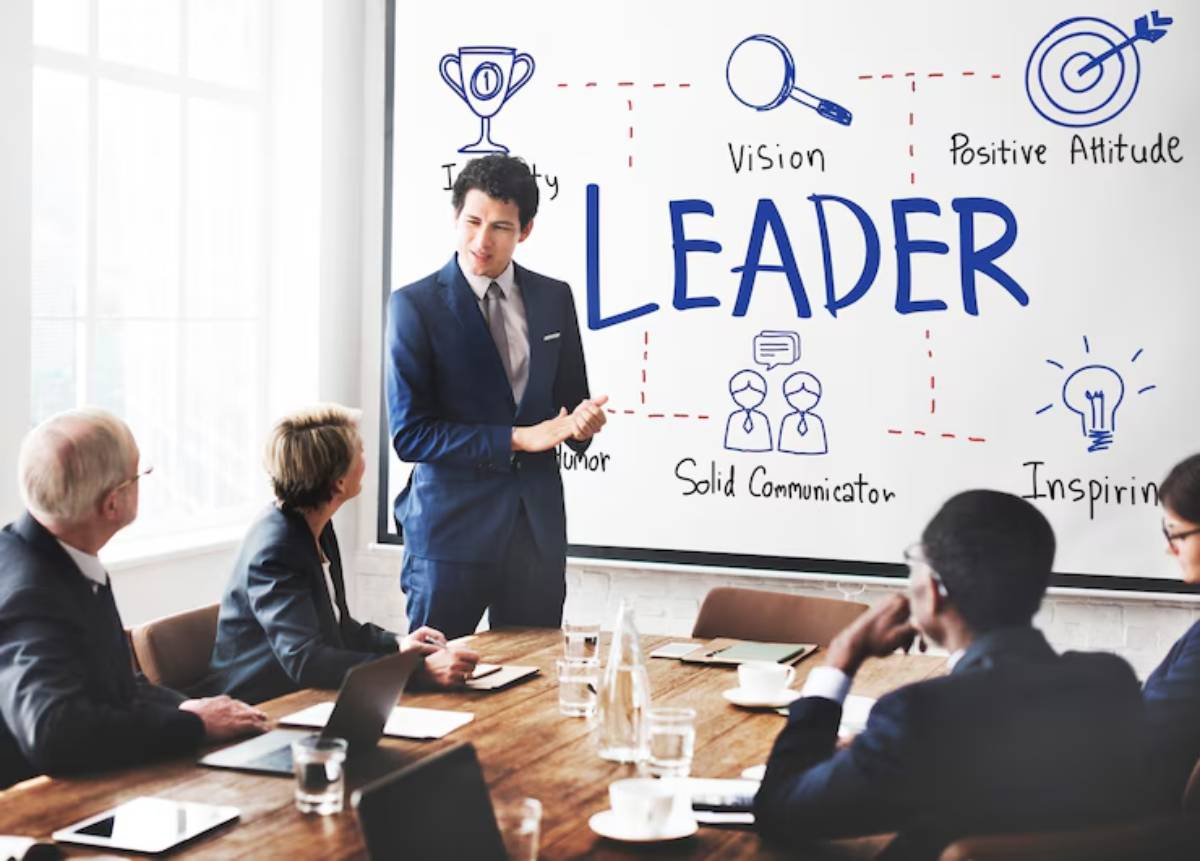 A business meeting with participants listening to a speaker presenting leadership qualities on a whiteboard: "LEADER," "Vision," and "Positive Attitude."