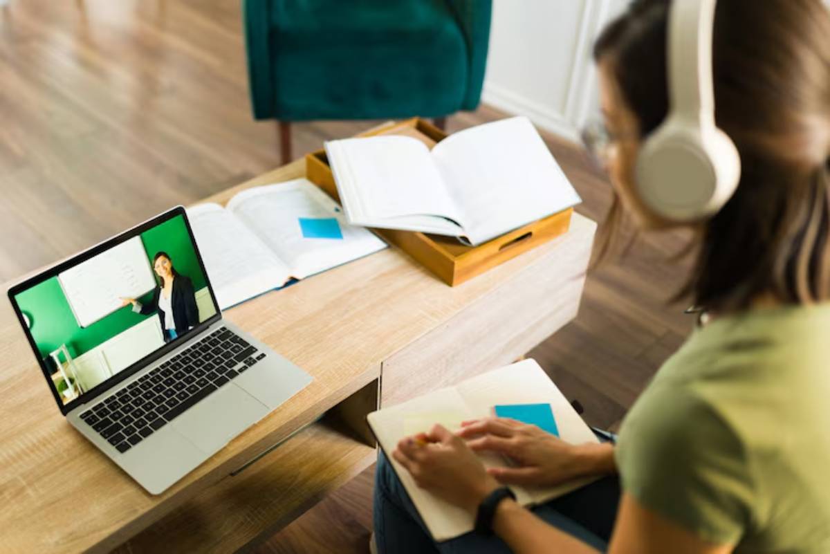 A person with headphones attends an online class, taking notes beside a laptop and open books on a coffee table