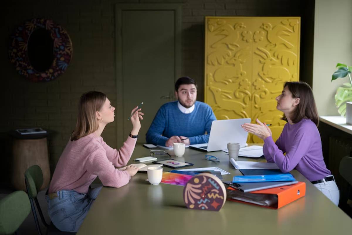 A group of three people engaged in discussion around a table with a laptop, notebooks, and coffee cups; colorful decor in the background.