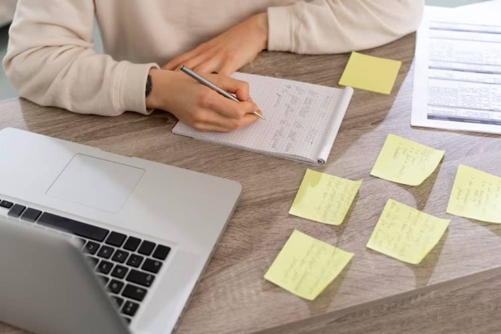 A person writes notes in a notepad next to a laptop, surrounded by yellow sticky notes, illustrating work session.